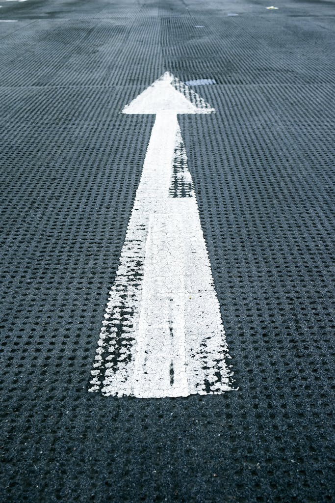 Close-up of a white directional arrow on a textured asphalt road, symbolizing forward movement and guidance.