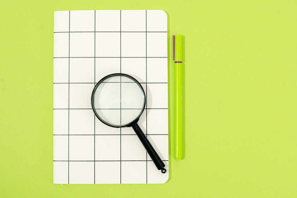 Flat lay of notebook, magnifying glass, and pen on a green background, perfect for office themes.