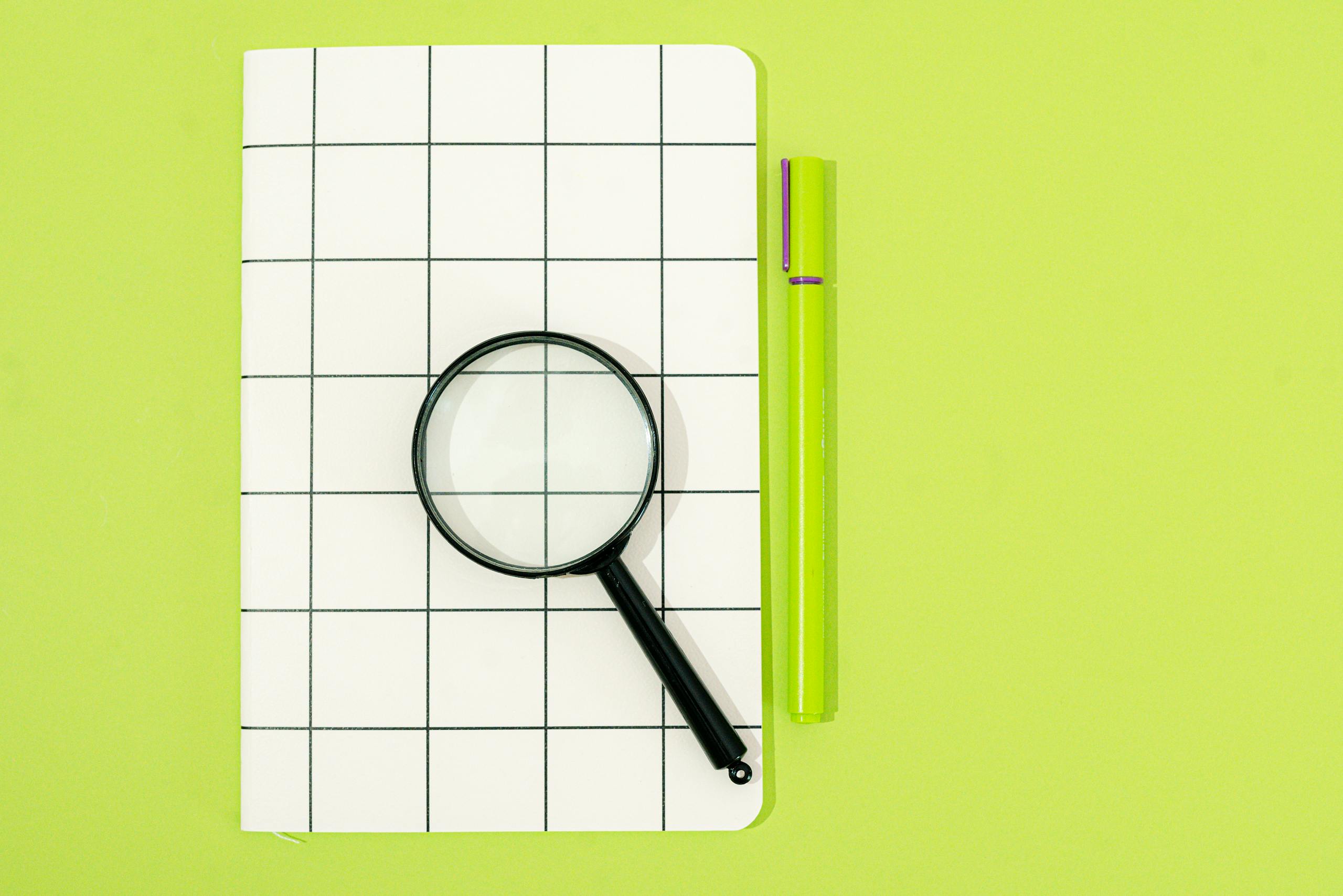 Flat lay of notebook, magnifying glass, and pen on a green background, perfect for office themes.