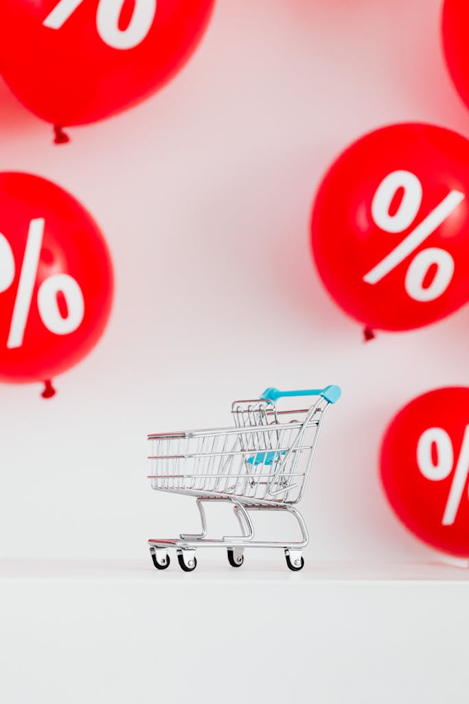 Mini shopping cart with red percentage balloons on a white background, symbolizing sales.