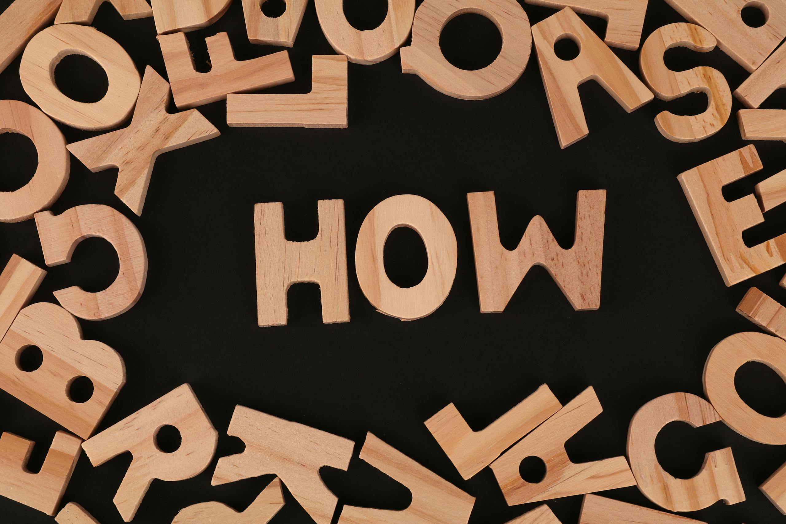 Top view of scattered wooden letters with 'HOW' centered on a dark surface, promoting inquiry.