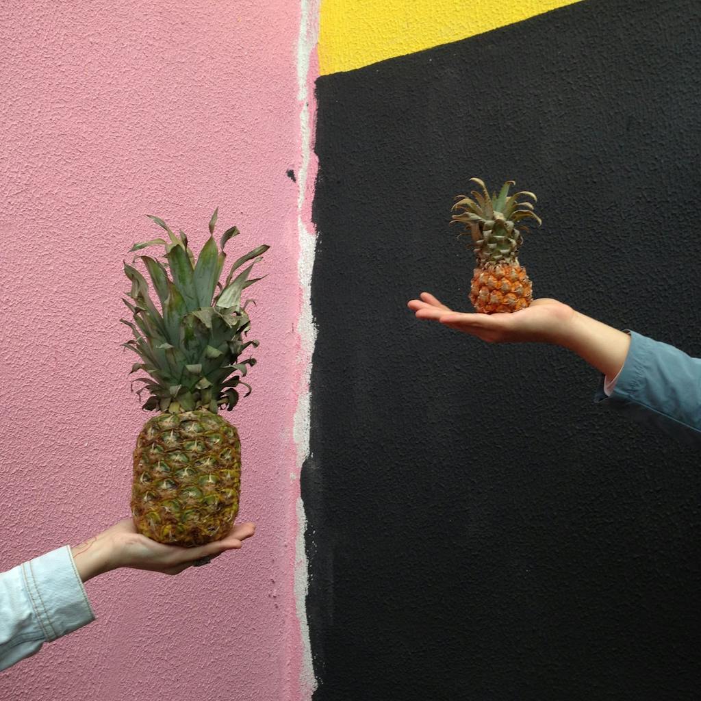 Two hands hold different sized pineapples against a colorful textured wall, showcasing a size contrast theme.